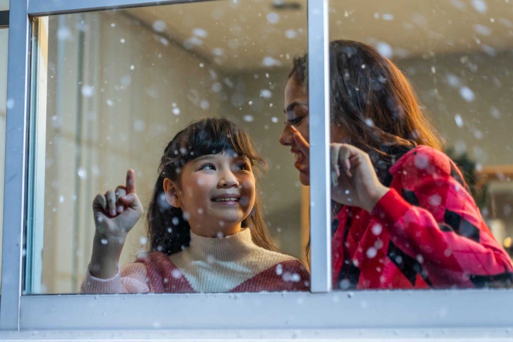 A mother and daughter looking out a window at falling snow. This is here to represent the question, can you replace windows in winter?